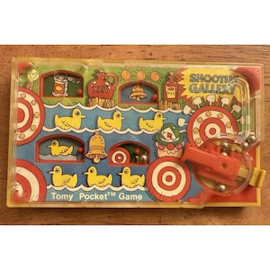 Puede incluir: Un juego de bolsillo Tomy vintage, Shooting Gallery, en una caja de plástico transparente con una ilustración colorida. El juego incluye patos amarillos, dianas y una pistola roja y amarilla. El texto "SHOOTING GALLERY" es visible.