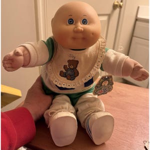 Könnte beinhalten: Eine Cabbage Patch Kids Puppe mit Glatze, blauen Augen und Grübchen in den Wangen. Die Puppe trägt ein grün-weißes Langarmshirt, eine weiße Hose und weiße Schuhe. Ein cremefarbener Lätzchen zeigt eine Grafik eines braunen Teddybären und den Text Sly.