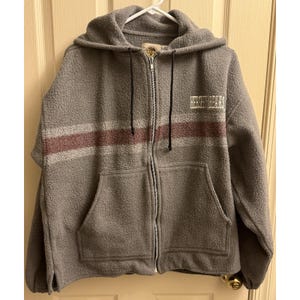 May include: A gray fleece zip-up hoodie with a hood and two front pockets. The hoodie features a horizontal stripe in burgundy and light gray across the chest. The words "HERSHEY BEARS" are printed in white on the right chest.