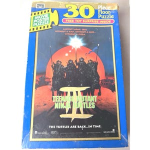 May include: A 30-piece floor puzzle featuring the Teenage Mutant Ninja Turtles II movie poster. The box is blue with yellow and red text, and the poster art depicts the turtles in ancient Japan. Includes a free toy surprise.