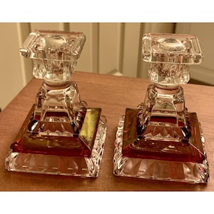 Puede incluir: Dos candelabros de vidrio transparente con base y parte superior cuadradas. La base tiene un tinte rojo y la parte superior una abertura cuadrada para una vela. Los candelabros están sobre una superficie de madera.