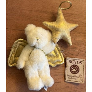 Könnte beinhalten: Ein cremefarbener Engelbär mit goldenen Flügeln und einem goldenen Sternornament. Der Bär hat einen goldenen Heiligenschein und hält einen goldenen Stern. Ein Etikett von The Head Bean Collection ist angebracht.