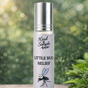 Puede incluir: Una botella de vidrio transparente con tapa plateada, etiquetada "Kind & Simple LITTLE BUG RELIEF". La etiqueta presenta un dibujo de mosquito y el texto "10ML". La botella está sobre un fondo verde borroso.