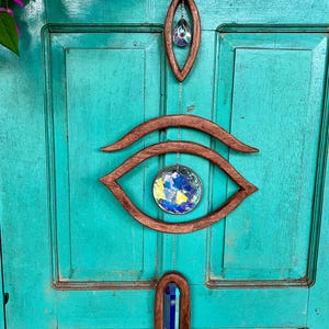 May include: A wooden eye-shaped wall hanging with iridescent glass crystals. The hanging features a large eye shape with a smaller crystal in the centre, and two smaller crystal drops hanging below.