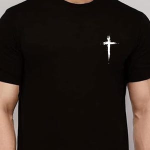 May include: Black crew neck t-shirt with a white cross graphic on the left chest. The cross has a textured, brushstroke appearance. The shirt is a solid black color and appears to be made of a soft material.