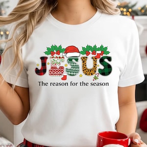 May include: White t-shirt with the word "JESUS" in festive Christmas patterns and a Santa hat above the letter "S". The text "The reason for the season" is below the word. The person is holding a Santa mug.
