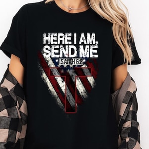 May include: Black t-shirt featuring the text "HERE I AM, SEND ME" above a distressed American flag design with a cross. The text "ISAIAH 6:8" is below the flag. The shirt has short sleeves.