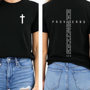 May include: Black crew neck t-shirt with a white cross graphic on the front. The back of the shirt features the word "PROVERBS" and a vertical text with a bible verse. The shirt is paired with blue jeans.