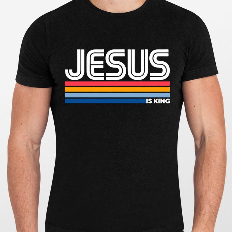 K&ouml;nnte beinhalten: Schwarzes T-Shirt mit dem Wort "JESUS" in wei&szlig;en, umrandeten Buchstaben &uuml;ber horizontalen Streifen in Rot, Orange, Blau und Marine. Der Text "IS KING" ist darunter in Wei&szlig; gedruckt.