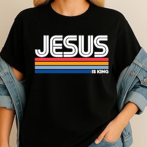 K&ouml;nnte beinhalten: Schwarzes T-Shirt mit dem Text "JESUS IS KING" in wei&szlig;en, umrandeten Buchstaben. Der Text befindet sich &uuml;ber einem horizontalen Streifendesign in Rot, Orange, Gelb und Blau.