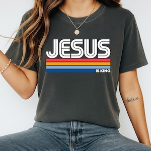 K&ouml;nnte beinhalten: Dunkelgraues T-Shirt mit dem Text "JESUS IS KING" in wei&szlig;en Blockbuchstaben &uuml;ber einem horizontalen Streifendesign in Rot, Orange, Gelb und Blau. Das Shirt hat einen Rundhalsausschnitt und kurze &Auml;rmel.