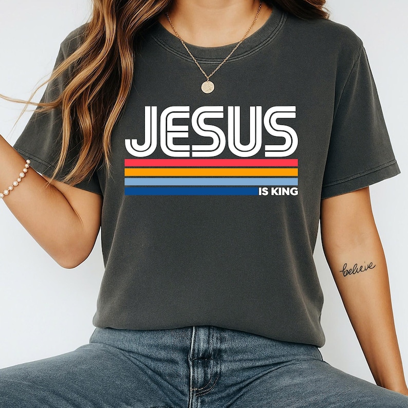 K&ouml;nnte beinhalten: Dunkelgraues T-Shirt mit dem Text "JESUS IS KING" in wei&szlig;en Blockbuchstaben &uuml;ber einem horizontalen Streifendesign in Rot, Orange, Gelb und Blau. Das Shirt hat einen Rundhalsausschnitt und kurze &Auml;rmel.