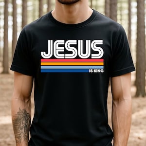 K&ouml;nnte beinhalten: Schwarzes T-Shirt mit dem wei&szlig;en Text "JESUS IS KING" &uuml;ber einem horizontalen Streifendesign in Rot, Orange, Gelb und Blau. Das Shirt hat einen Rundhalsausschnitt.