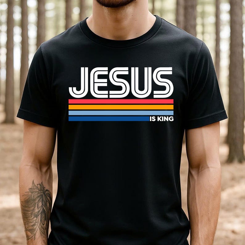 K&ouml;nnte beinhalten: Schwarzes T-Shirt mit dem wei&szlig;en Text "JESUS IS KING" &uuml;ber einem horizontalen Streifendesign in Rot, Orange, Gelb und Blau. Das Shirt hat einen Rundhalsausschnitt.