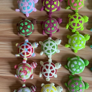 Puede incluir: Una colección de dieciocho pequeñas tortugas tejidas a mano en crochet, dispuestas en tres columnas verticales. Cada tortuga presenta una combinación de colores distinta, incluyendo tonos de rosa, verde, amarillo, rojo, blanco y marrón.
