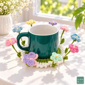 Puede incluir: Una taza de cerámica de color verde azulado oscuro sobre un posavasos de ganchillo redondo, verde, blanco y multicolor. El posavasos presenta varias flores de ganchillo tridimensionales pequeñas en tonos rosa, azul, amarillo, morado y verde claro.
