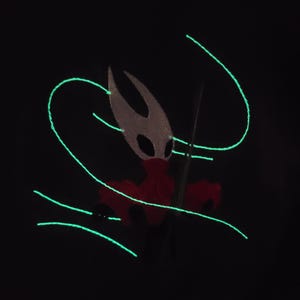 May include: A black background featuring a glowing green design of curved lines and a character with a white head, red body, and a black spear. The character has a mask with black eyes.