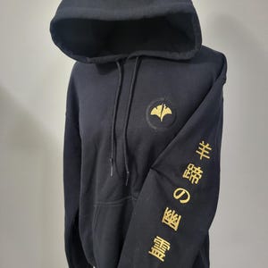 May include: Black hooded sweatshirt with a gold emblem on the chest and gold Japanese text on the sleeve. The sweatshirt has a drawstring hood and a front pocket. The design features a stylized leaf.