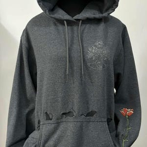 May include: Dark gray hoodie with a drawstring hood and a kangaroo pocket. Features a distressed design at the pocket and a floral embroidery on the sleeve. A tree and moon design is embroidered on the chest.