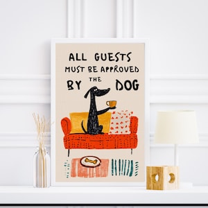 Puede incluir: Una impresión artística enmarcada con el texto "ALL GUESTS MUST BE APPROVED BY THE DOG". La ilustración presenta un perro negro en un sofá rojo sosteniendo una taza. La impresión tiene un fondo neutro y está decorada con un hueso y una alfombra.