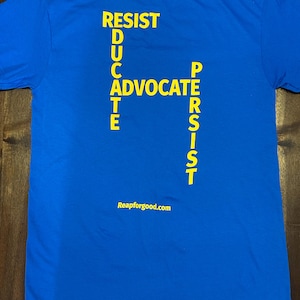 May include: A royal blue t-shirt with the words "RESIST", "EDUCATE", "ADVOCATE", and "PERSIST" printed in yellow, forming a cross. The words "Reapforgood.com" are printed in yellow at the bottom of the shirt.