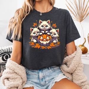 May include: Dark gray t-shirt featuring a Halloween design. The graphic shows three fluffy white kittens with blue eyes, nestled around a carved orange pumpkin. Autumn leaves and stars surround the kittens and pumpkin.