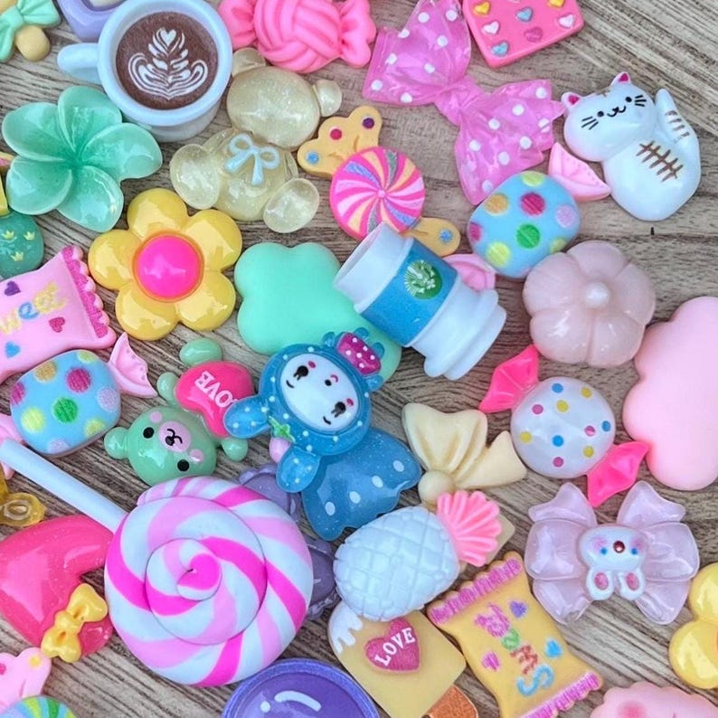 May include: A collection of colorful resin embellishments, including flowers, candies, bows, and a coffee cup. The assortment features various shapes and colors, such as pink, blue, yellow, and white, with some pieces displaying polka dots and other patterns.