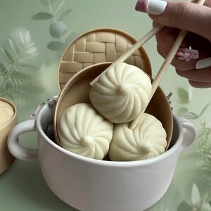 May include: Three white, spiraled, dumpling-shaped objects are nestled inside a small, round, tan-coloured bamboo steamer basket. The steamer is placed within a white ceramic bowl with two side handles, while wooden chopsticks hold one dumpling.