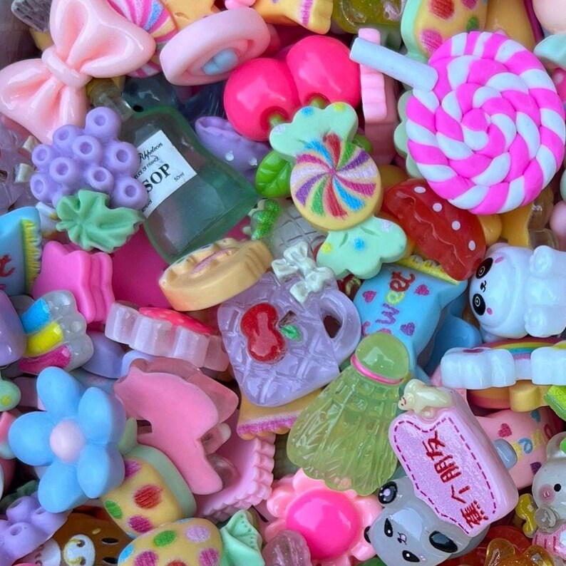 May include: A colorful assortment of miniature resin charms in various shapes, including bows, candies, flowers, and a bottle with the text "SOP". The charms are in shades of pink, purple, green, and yellow, creating a playful and whimsical aesthetic. A variety of sweets and treats.