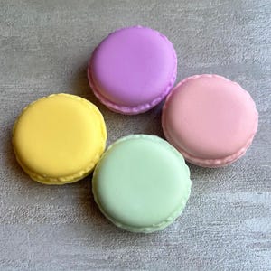 Może przedstawiać: Cztery kolorowe makaroniki w pastelowych odcieniach żółtego, fioletowego, różowego i zielonego ułożone są na szarej powierzchni. Każdy makaronik ma gładką górę i pofałdowany brzeg, prezentując klasyczny design deseru.