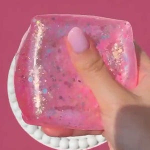 May include: A square, translucent pink slime toy filled with multicolored glitter and small iridescent sequins. The texture is gelatinous and pliable, showing a vibrant pink hue throughout the material with light reflecting off the embedded sparkles.