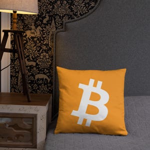 May include: A yellow throw pillow with a white Bitcoin symbol on it. The pillow is sitting on a gray couch.