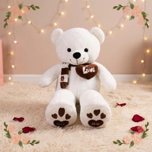 Pode incluir: Um urso de peluche branco com um cachecol às riscas castanhas e brancas, um emblema castanho em forma de coração com a palavra Love bordada a branco e detalhes de patas castanhas nos pés com as palavras love e you nas solas.