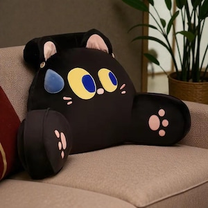 May include: A black cat-shaped plush pillow with yellow and blue eyes, pink paw prints, and a blue teardrop. The pillow has a headrest and armrests, and is designed to be used as a backrest. The pillow is on a beige couch.