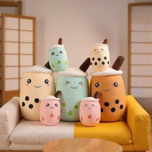 Puede incluir: Una colección de cojines de peluche con forma de bebida de té de burbujas en varios colores pastel, incluyendo amarillo, rosa y verde. Cada cojín tiene una cara sonriente y una pajita marrón. Algunos tienen bobas negras y diseños de frutas.