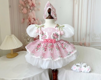 May include: A pink and white baby dress with matching bonnet and shoes. The dress features floral embroidery, lace trim, and puffy sleeves. The bonnet and shoes are also decorated with floral accents. The dress is displayed on a mannequin.