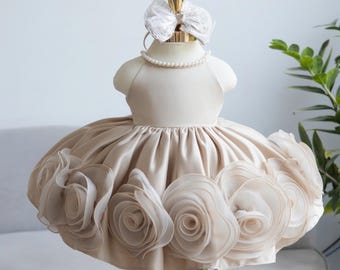 May include: A beige satin dress with a full skirt adorned with large, layered fabric roses. The dress is displayed on a mannequin with a pearl necklace and a white lace bow headband. The dress is designed for a baby or toddler.