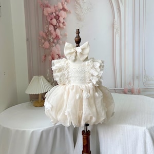 Peut inclure: Robe de bébé ivoire avec un grand nœud, des détails en dentelle et des accents floraux. La robe est présentée sur un mannequin en bois, mettant en valeur ses manches à volants et sa jupe superposée. Idéale pour une occasion spéciale.