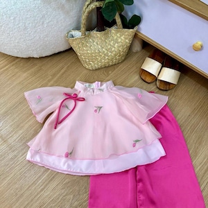 May include: A child's two-piece pink outfit. The top has a mandarin collar, short sleeves, and embroidered floral details. The trousers are a solid, vibrant pink. The outfit is made of a lightweight fabric, possibly silk. The outfit is suitable for a child.