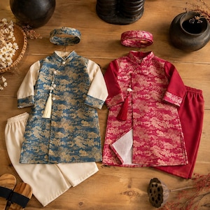 Puede incluir: Dos conjuntos de ropa de estilo tradicional, uno azul y otro rojo, cada uno con una túnica de manga larga con un patrón de paisaje dorado, una diadema a juego y pantalones de color sólido. Las túnicas tienen cierres laterales con borlas.
