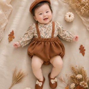 May include: A baby in a brown beret, floral print top, and brown suspender shorts. The outfit is completed with brown shoes with cream bows. The scene is decorated with pumpkins and leaves, evoking an autumnal theme.