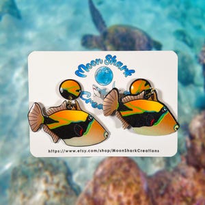 Handmade rectangular triggerfish earrings crafted from shrink plastic with a glossy UV resin front. These lightweight dangle earrings feature bold rectangular triggerfish with striking black bodies, bright yellow markings, and electric blue accents inspired by tropical reef life. Eye-catching and unique, they add a vibrant ocean vibe—perfect for marine life lovers and statement jewelry fans. By Moon Shark Creations.