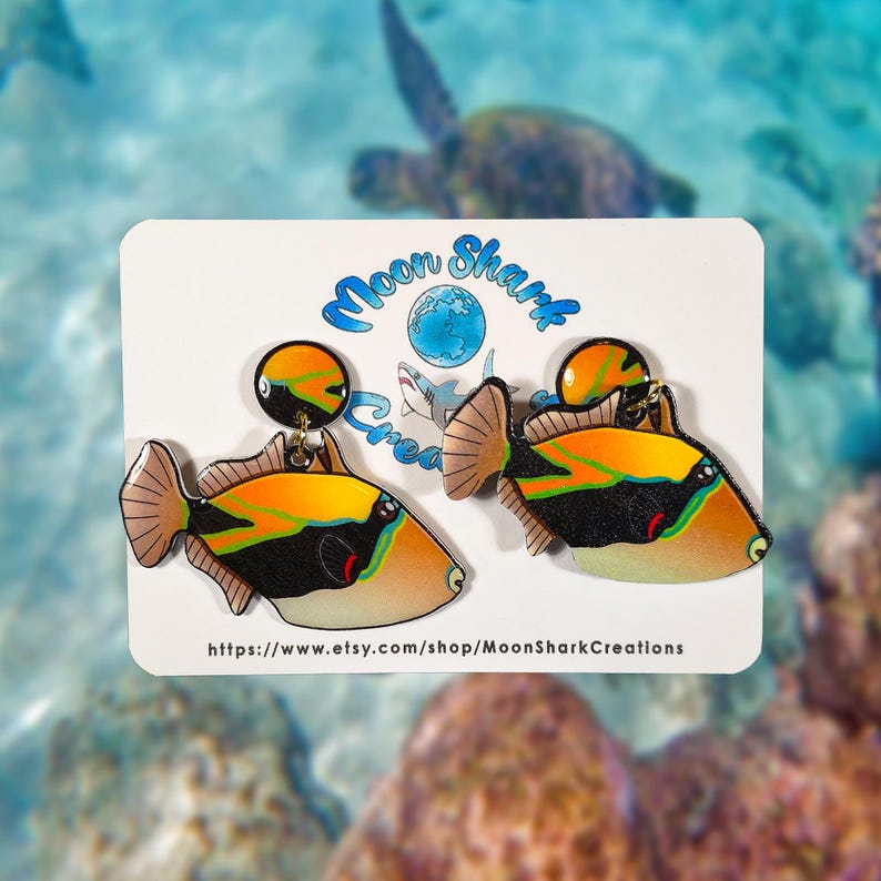 Handmade rectangular triggerfish earrings crafted from shrink plastic with a glossy UV resin front. These lightweight dangle earrings feature bold rectangular triggerfish with striking black bodies, bright yellow markings, and electric blue accents inspired by tropical reef life. Eye-catching and unique, they add a vibrant ocean vibe—perfect for marine life lovers and statement jewelry fans. By Moon Shark Creations.