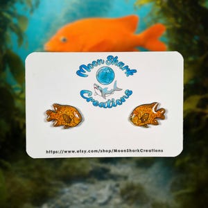 May include: A pair of orange fish-shaped earrings with glitter accents are displayed on a white card. The card features the "Moon Shark Creations" logo and a website address. An out-of-focus orange fish is in the background.