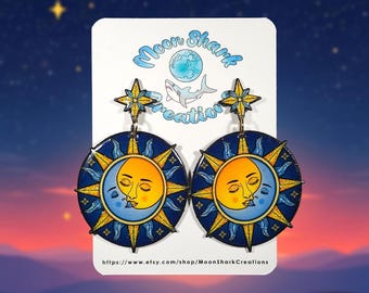 May include: A pair of celestial-themed earrings. Each earring features a circular design with a yellow sun, a blue moon with a face, and a dark blue background with yellow star accents. The earrings are suspended from a star-shaped top.