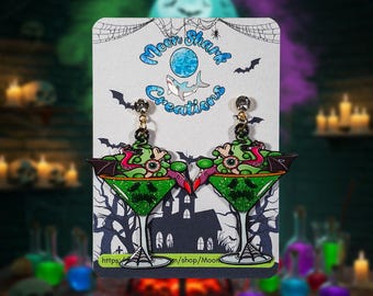 Peut inclure: Une paire de boucles d'oreilles sur le thème d'Halloween. Chaque boucle d'oreille présente un design de verre à martini vert avec un visage effrayant, surmonté d'une créature verte, rose et noire. Les boucles d'oreilles sont sur une carte avec le texte "Moon Shark Creations".