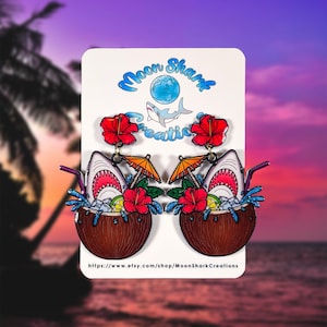 Op de afbeelding: Nieuwe oorbellen met een tropisch thema. Elke oorbel heeft een haaienkop die uit een kokosdrank komt, compleet met een rietje, limoen en hibiscusbloem. De oorbellen hebben een blauw en wit glittereffect. De kaart leest "Moon Shark Creations".