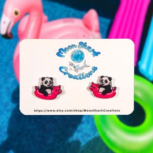 May include: A pair of panda bear earrings. Each earring features a black and white panda riding a pink inflatable pig. The earrings are displayed on a white card with a blue logo and the text "Moon Shark Creations".