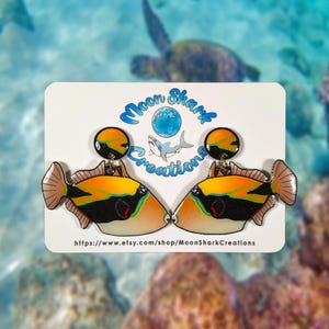 May include: A pair of colorful fish-shaped earrings with orange, yellow, black, and white details. The earrings are on a white card with the text "Moon Shark Creations" and a website address. The background shows an underwater scene.