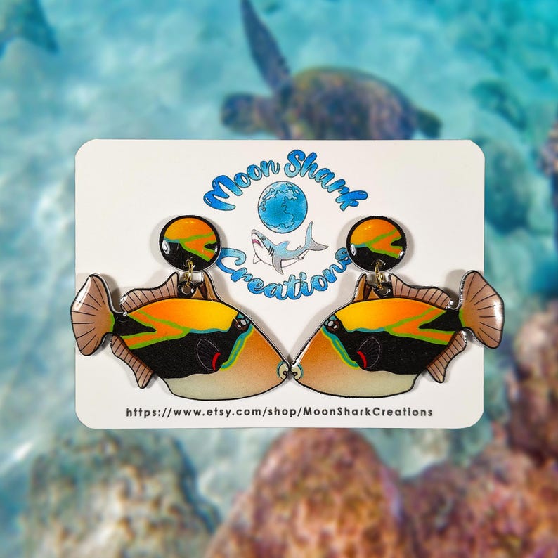 Handmade rectangular triggerfish earrings crafted from shrink plastic with a glossy UV resin front. These lightweight dangle earrings feature bold rectangular triggerfish with striking black bodies, bright yellow markings, and electric blue accents inspired by tropical reef life. Eye-catching and unique, they add a vibrant ocean vibe—perfect for marine life lovers and statement jewelry fans. By Moon Shark Creations.